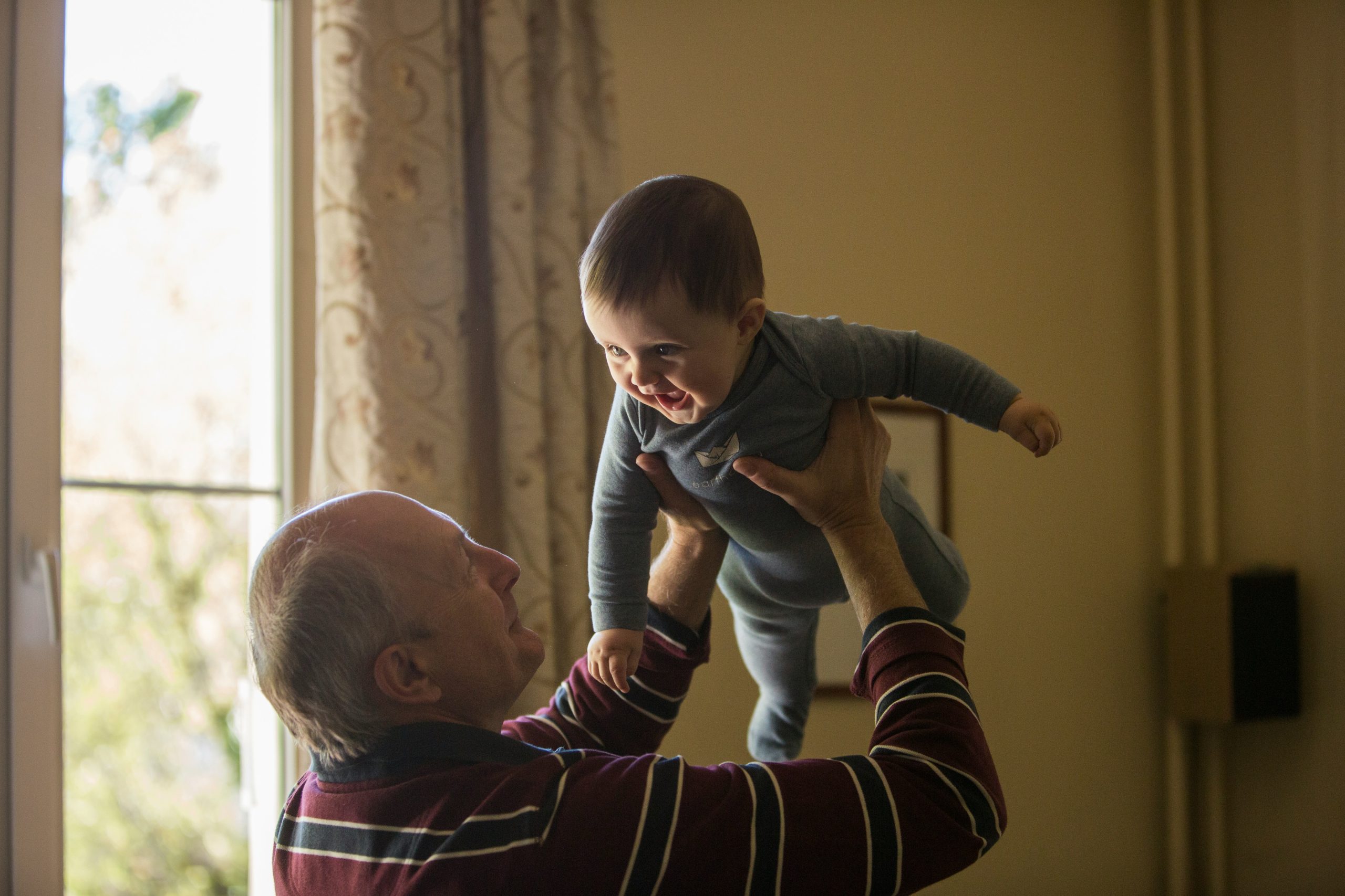 man-wearing-maroon-white-and-blue-stripe-long-sleeved-shirt-lifting-up-baby-wearing-gray-onesie